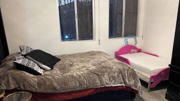 Een slaapkamer met een bed, een klein tafeltje, een roze stoel en twee ramen.