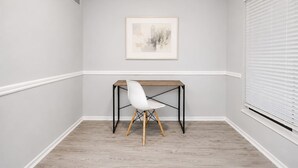 A corner room with a white chair, a wooden table, and a framed picture on the wall.