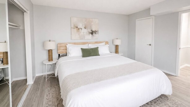 A bedroom with a large bed, two bedside tables with lamps, a wooden headboard, a wall-mounted abstract painting, and a wardrobe.