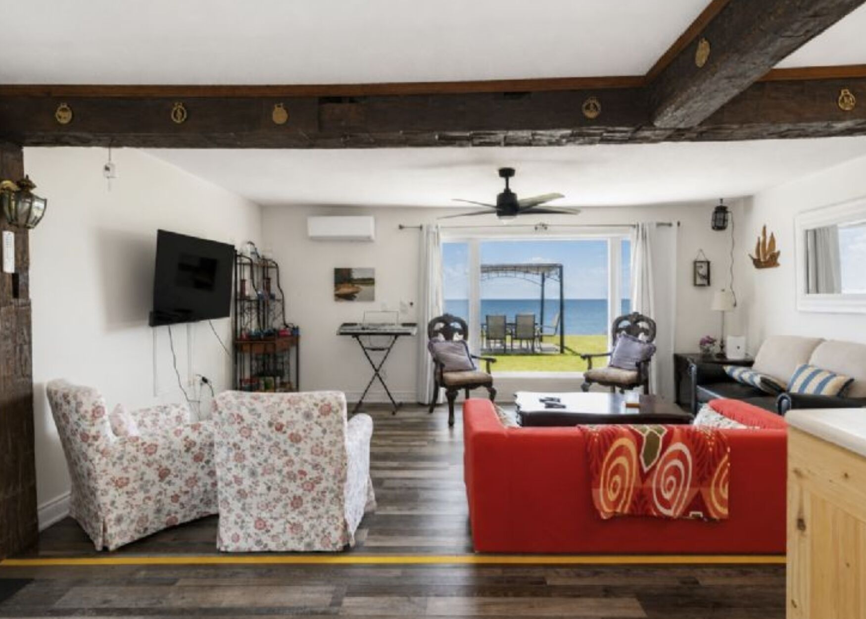 A living room with a red sofa, two armchairs, a television, and a view of the sea through large windows.