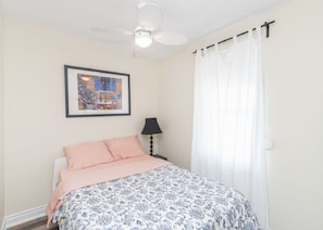 Un dormitorio con cama, ventilador de techo, cuadro enmarcado, lámpara y ventana con cortinas.