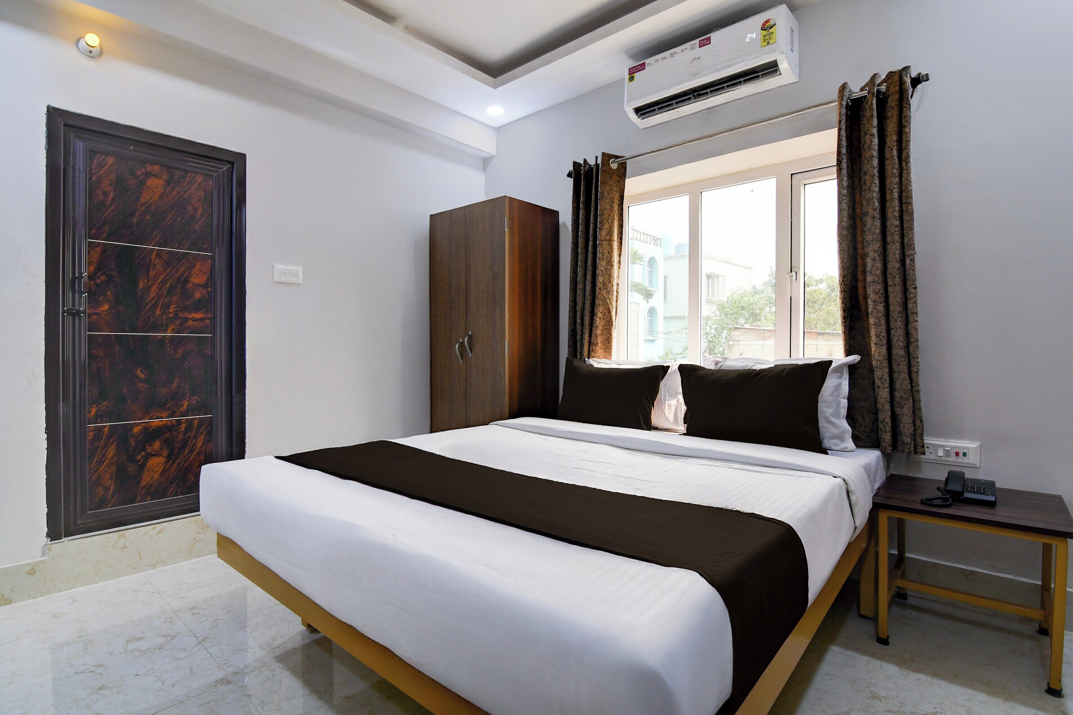A hotel room with a bed, a wardrobe, a nightstand, an air conditioning unit, and a window with curtains.