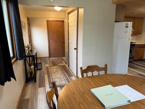 A room with a wooden dining table, chairs, a small rug, a white door, and a kitchen area with cabinets and a refrigerator.