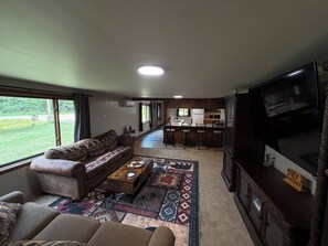 Ein Wohnzimmer mit einer Couch, einem Couchtisch und einem Fernseher. Es gibt eine Küchenzeile mit einer Bar und einem Esstisch.