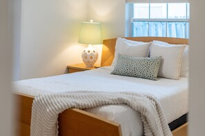 Un lit bien fait, avec une couette blanche et des oreillers, une tête de lit en bois et une lampe de chevet dotée d’un abat-jour à motifs.