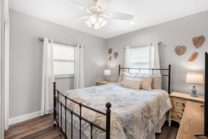 Une chambre à coucher comprenant un lit, deux tables de chevet, un ventilateur de plafond et des œuvres d’art décoratives murales.