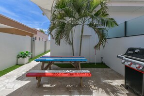 A patio with a barbecue grill, a table with a colorful top, and a palm tree. - Modern Aruba Studio · Stylish & Newly Fitted (Noord)
