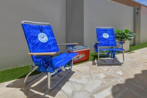 Duas cadeiras de praia com padrão de folhagem tropical e um logotipo, colocadas num pátio de pedra com uma planta em vaso por perto.
