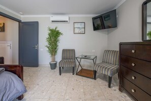 Ein kompakter Wohnraum mit Bett, Schreibtisch, zwei Stühlen, einem Schminktisch und einem Topf mit Pflanze, sowie einem Flachbildfernseher an der Wand.