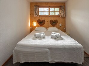 Un dormitorio con cabecera de madera, dos adornos con forma de corazón, una ventana con cortinas y toallas dobladas con cuidado sobre la cama.