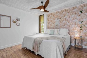 A bedroom with a bed, a ceiling fan, a bedside table with a lamp, and a wall with a leaf pattern. - Ma'alaea Ocean Front • Whales, Pool & Hot Tub (Wailuku)