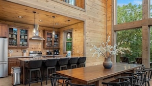 A modern kitchen with wooden cabinetry, a central island with bar stools, and a large window overlooking greenery. - 2 Cabin Retreat/Pool/2HotTubs/Playground/Sleeps 22 (Sevierville)