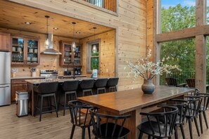 A modern kitchen with wooden cabinetry, a central island with bar stools, and a large window overlooking greenery. - 2 Cabin Retreat/Pool/2HotTubs/Playground/Sleeps 22 (Sevierville)