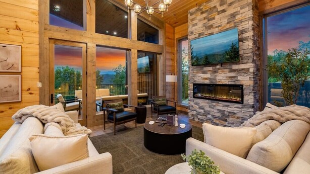 A spacious living room with a stone fireplace, comfortable seating, and large windows offering a view of the outdoors. - 2 Cabin Retreat/Pool/2HotTubs/Playground/Sleeps 22 (Sevierville)