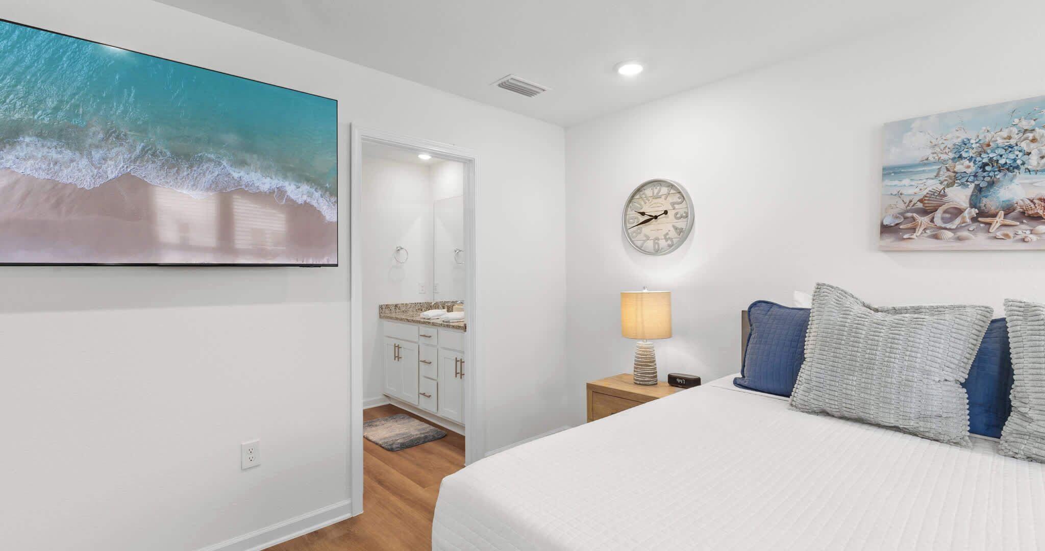 A bedroom with a bed, a nightstand, a lamp, a clock, and a painting of a beach scene.