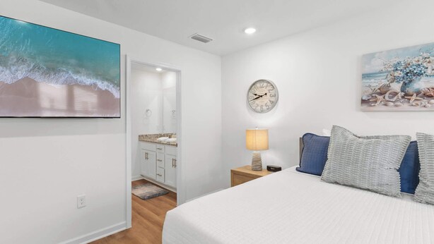 A bedroom with a bed, a nightstand, a lamp, a clock, and a painting of a beach scene. - Easy Days at Lost Bay - 3BDR (Orange Beach)