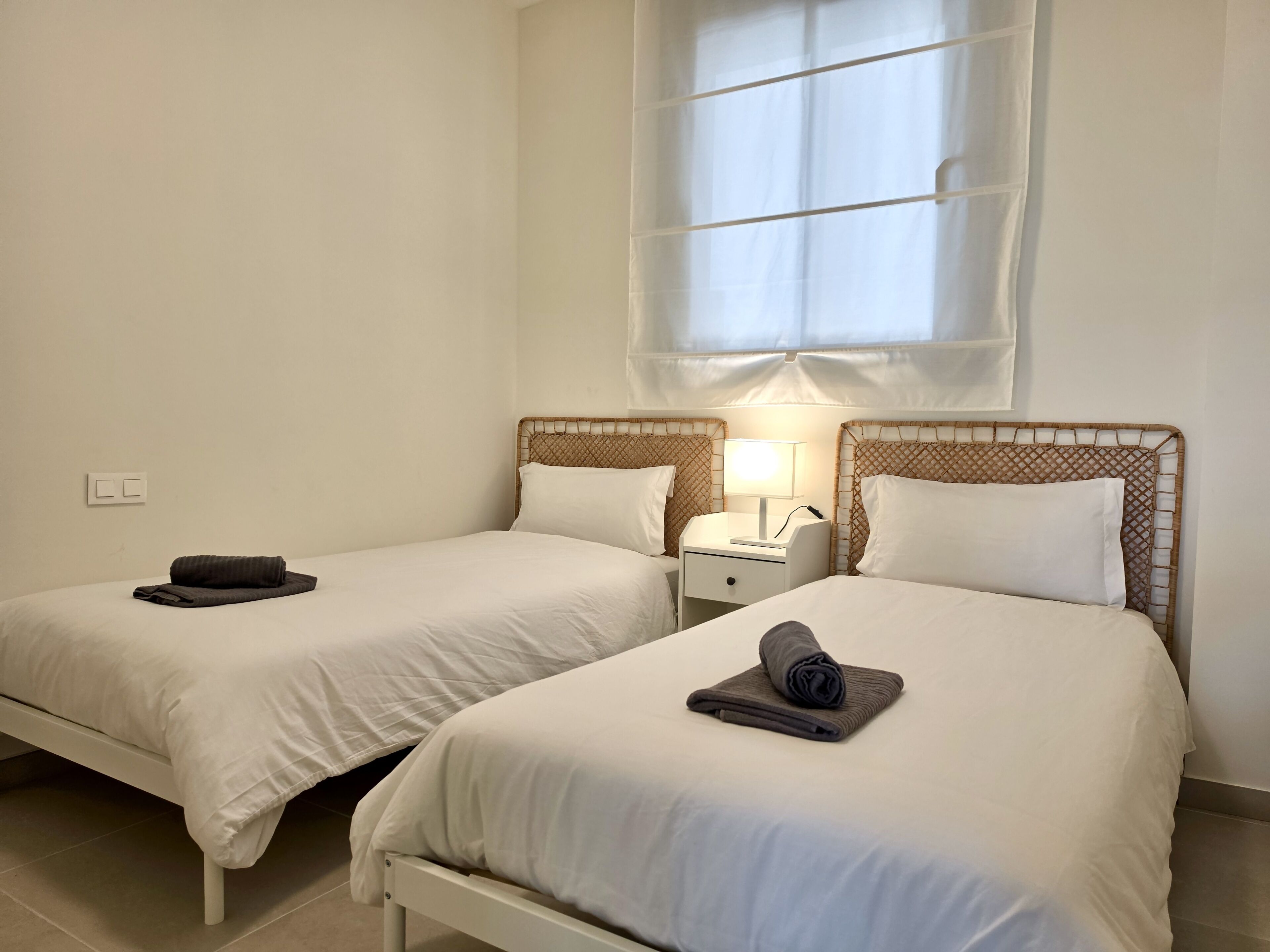 A hotel room with two single beds, each with a white mattress and matching pillows, a bedside table with a lamp, and a window above the beds.