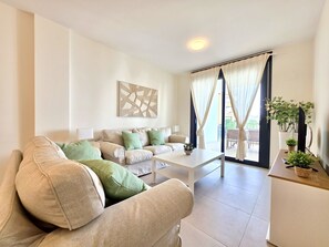 A modern living room with a sofa, a coffee table, and a view of the outdoors through large windows. - SR Calanova Golf and Beach 2-Bedroom Condo in Mijas Costa (Mijas)