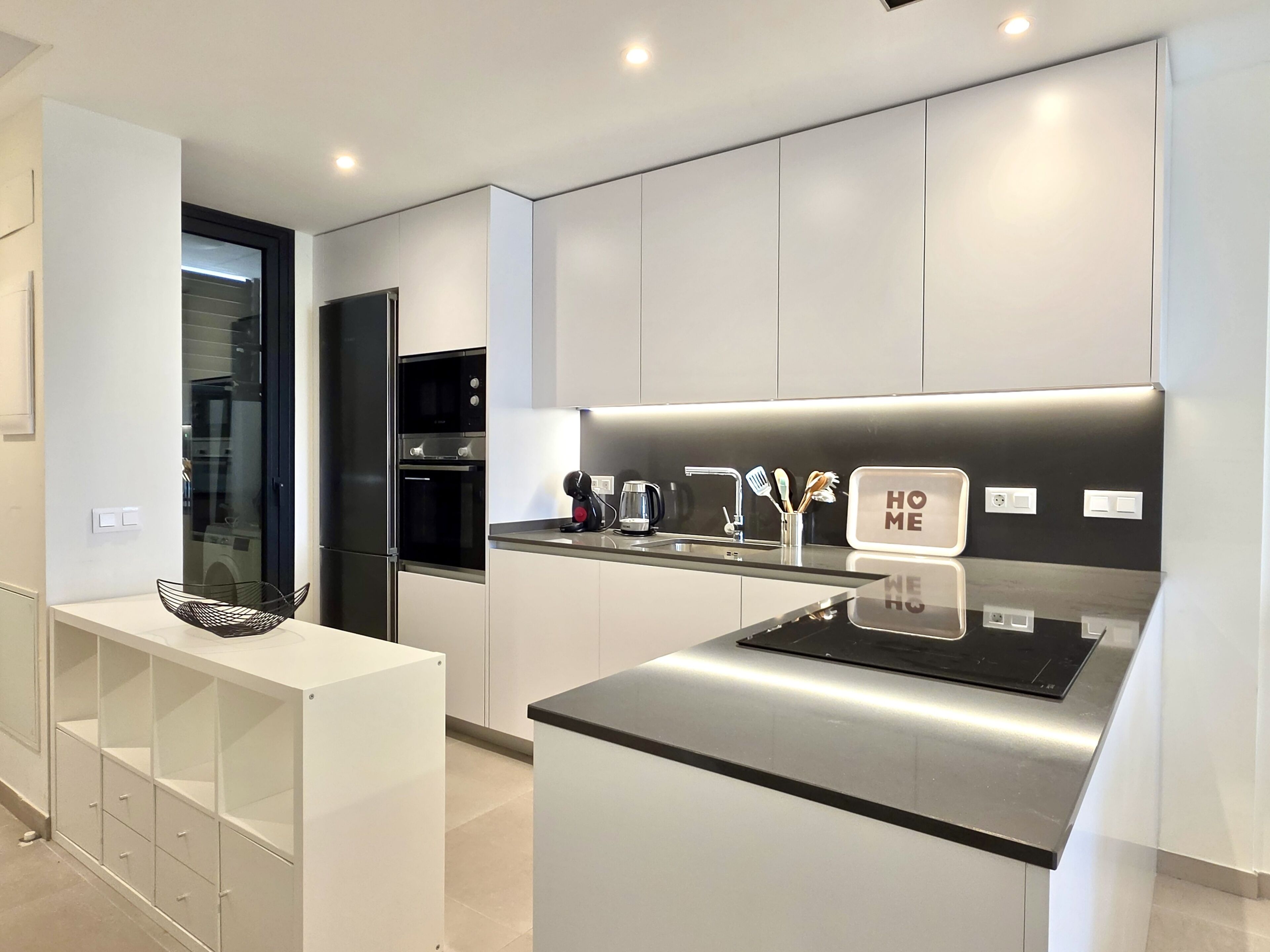 A modern kitchen with a built-in oven, a coffee maker, and a 'HOME' sign on the countertop.