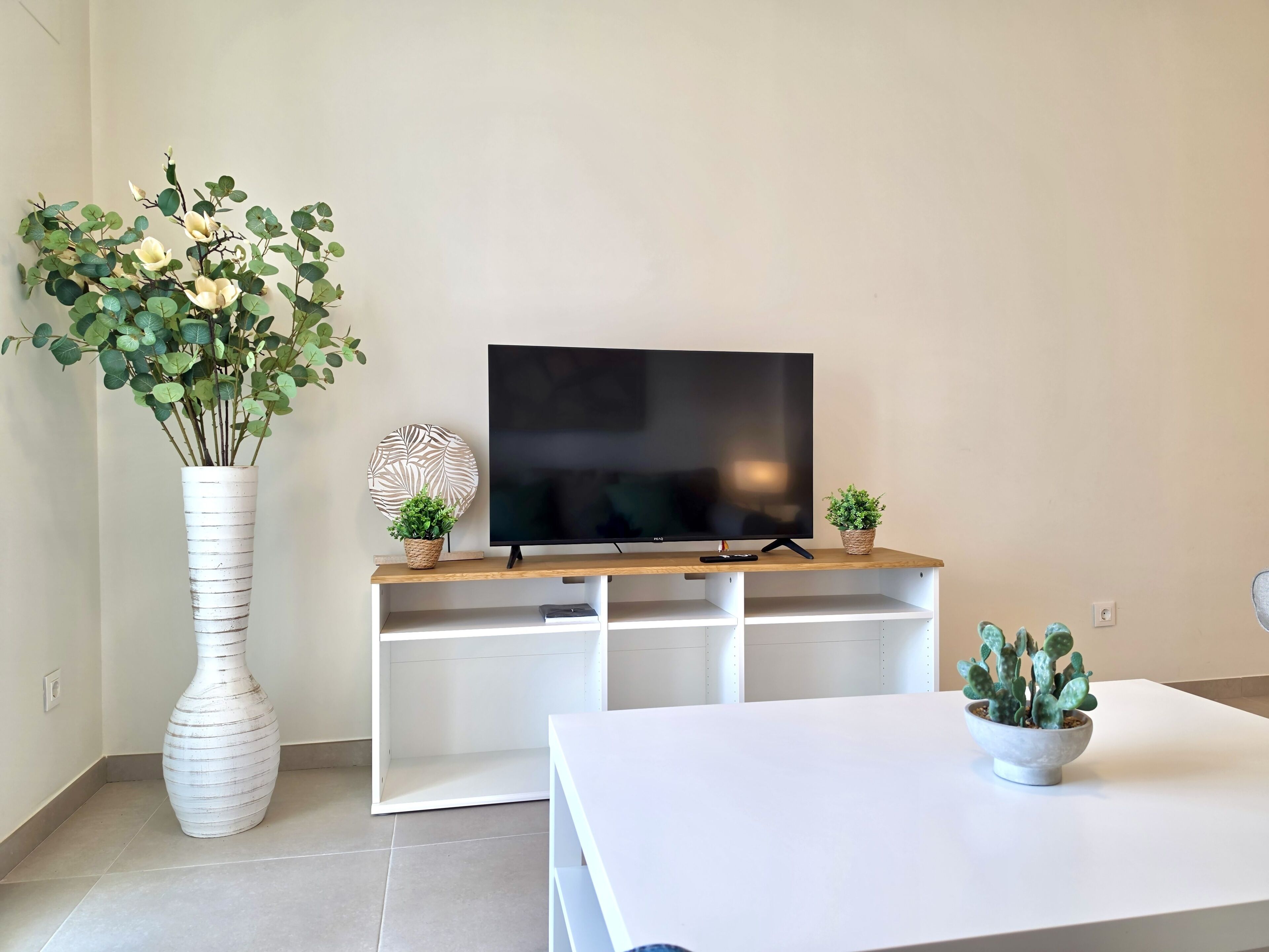 A living room with a flat-screen TV on a wooden stand, a white vase with flowers, and a small potted plant on a white table.