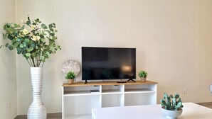 A living room with a flat-screen TV on a wooden stand, a white vase with flowers, and a small potted plant on a white table. - SR Calanova Golf and Beach 2-Bedroom Condo in Mijas Costa (Mijas)