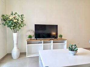 Ein Wohnzimmer mit einem Flachbildfernseher auf einem Holzboden, einer weißen Vase mit Blumen und einer kleinen Topfpflanze auf einem weißen Tisch.
