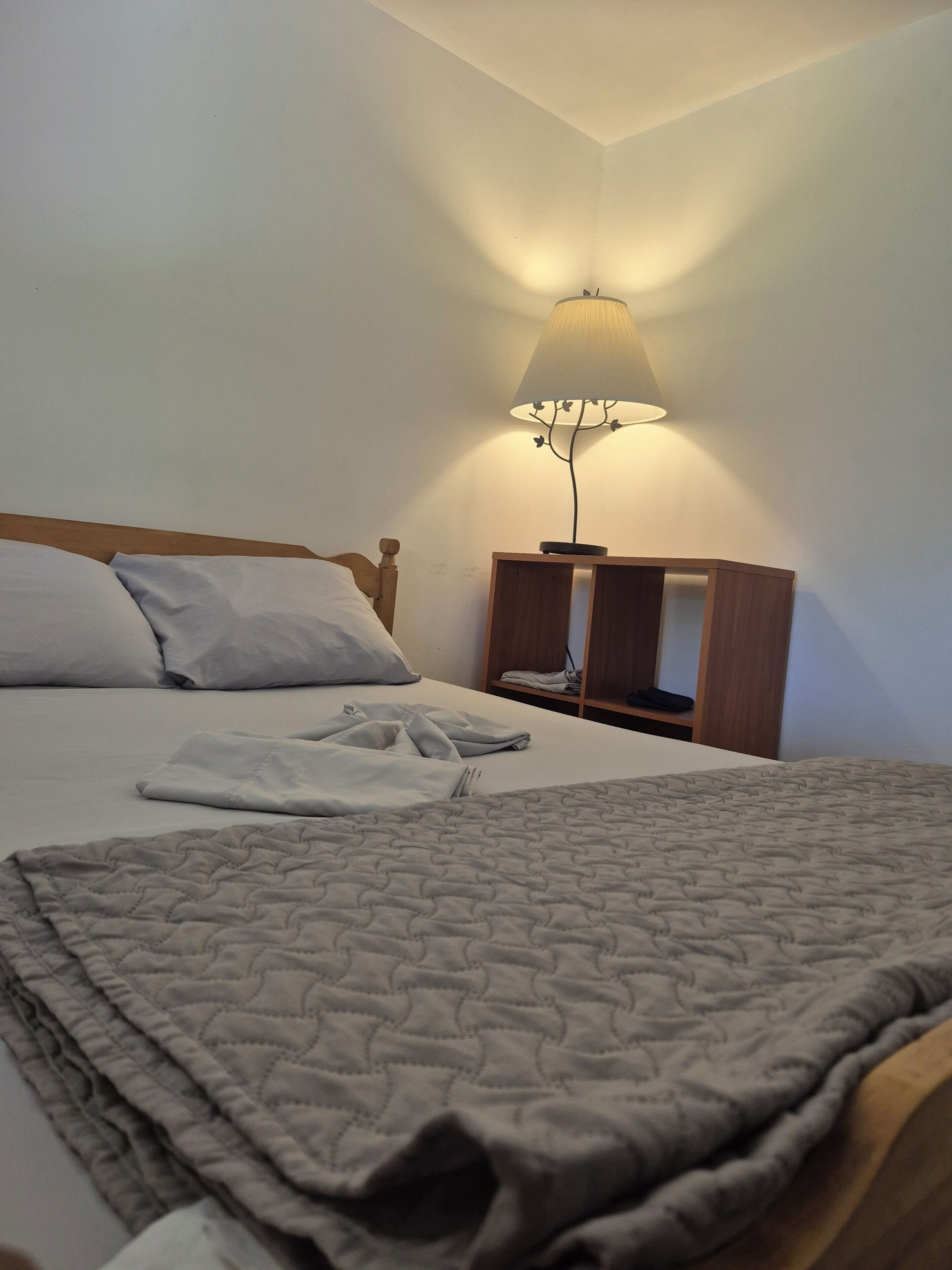 Un lit bien fait, recouvert d’une couverture matelassée, une table de chevet avec une lampe et une petite table en bois  à côté du lit.