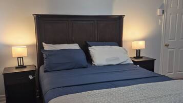 A bedroom with a dark wood headboard, two bedside tables with lamps, a bed with blue bedding, and a white door.