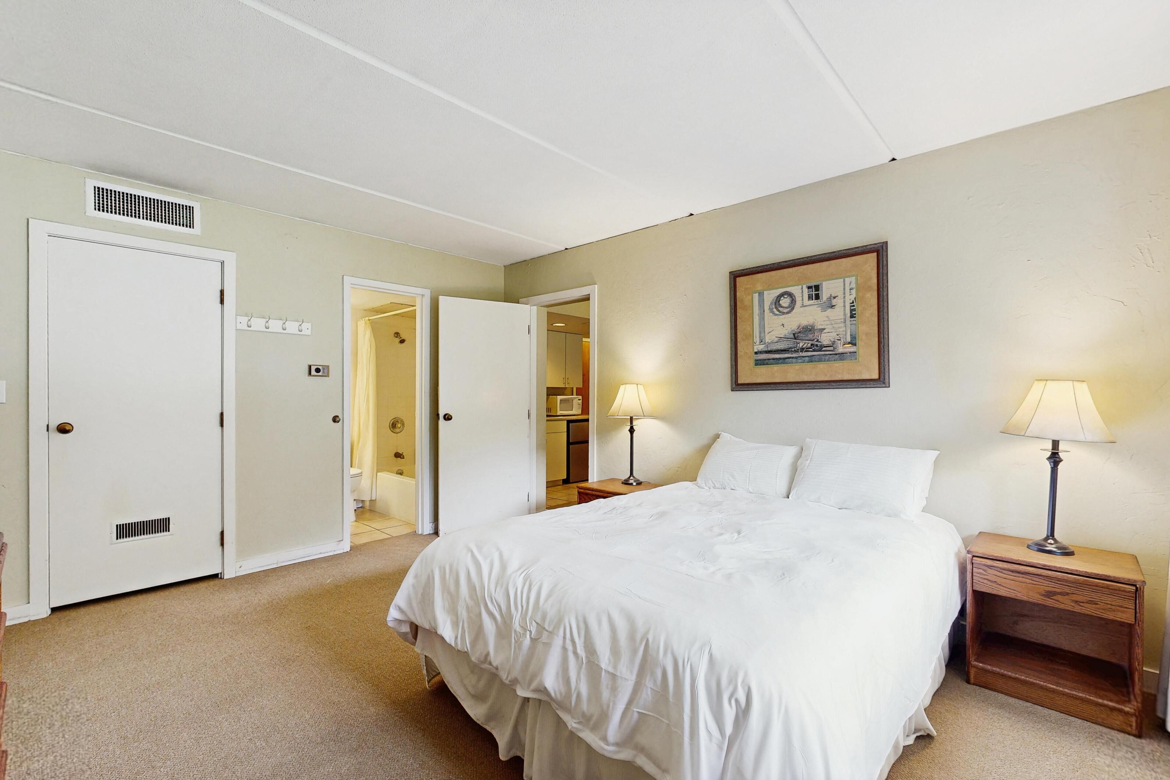 A hotel room with a bed, bedside lamps, a painting on the wall, and a door leading to a bathroom.
