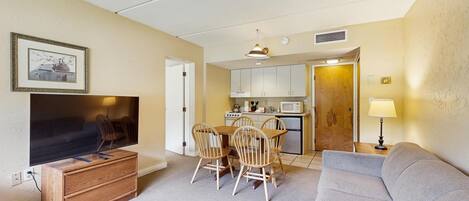 A compact living space with a television, a dining table with chairs, a kitchenette, and a sofa.