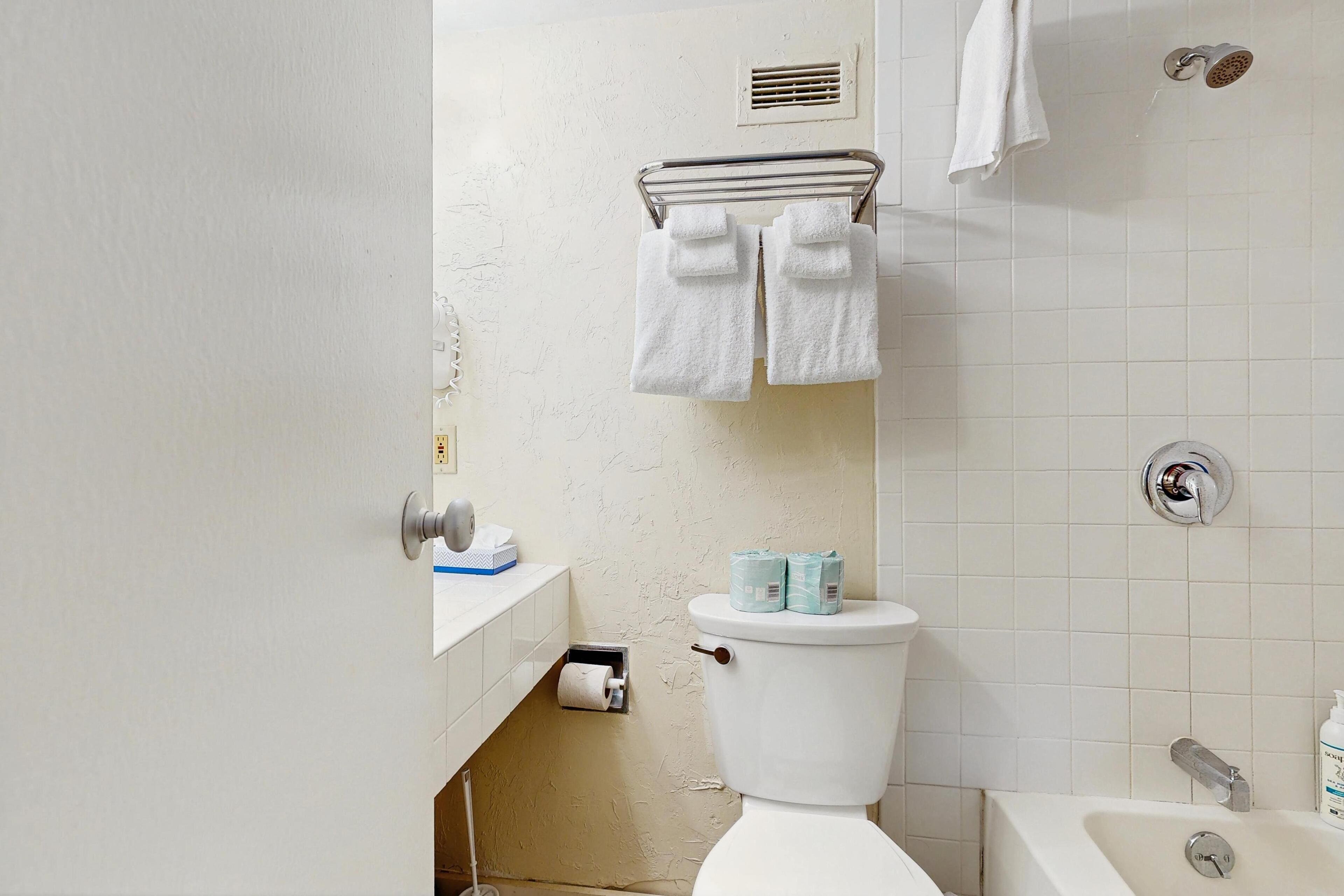 A bathroom with a toilet, a sink with a towel rack above, a roll of toilet paper, and a bottle of hand soap.