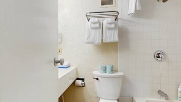 A bathroom with a toilet, a sink with a towel rack above, a roll of toilet paper, and a bottle of hand soap.