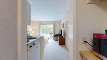 Un pasillo estrecho que conduce a un balcón con vistas a árboles, una kitchenette con estufa, una silla y un televisor.