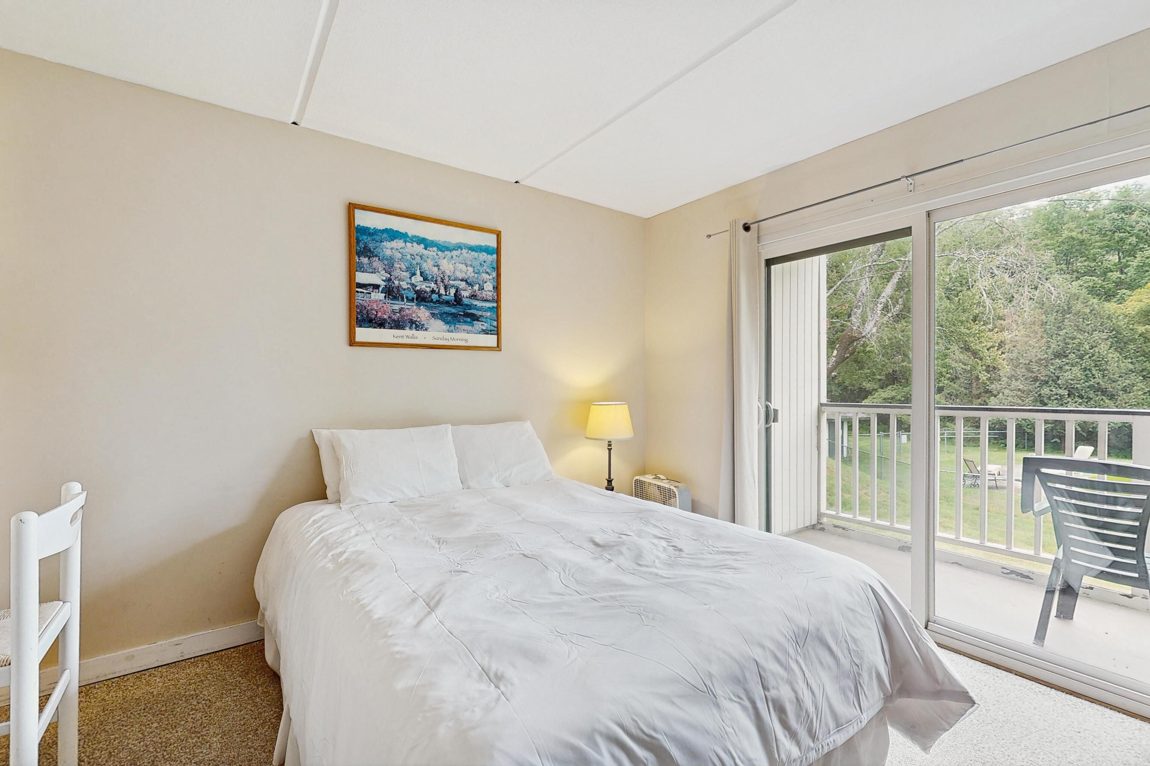 Habitación de hotel con cama, mesita de noche, lámpara y vistas al exterior a través de un ventanal corredero.