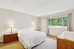A bedroom with a bed, two bedside lamps, a wooden nightstand, a window with curtains, and a view of greenery outside. - Sugarbush Condo: seasonal pool, ski, hike & golf (Warren)