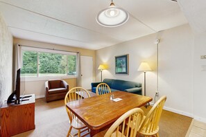 A room with a wooden dining table, chairs, a sofa, a television, and a window with a view of greenery. - Sugarbush-area condo with private queen bedroom and seasonal pool (Warren)