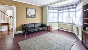 A spacious living room with a black leather sofa, a wooden floor, a large window with curtains, and a painting on the wall. - Ldn-hend in London With 3 Bedrooms and 1 Bathrooms (London)