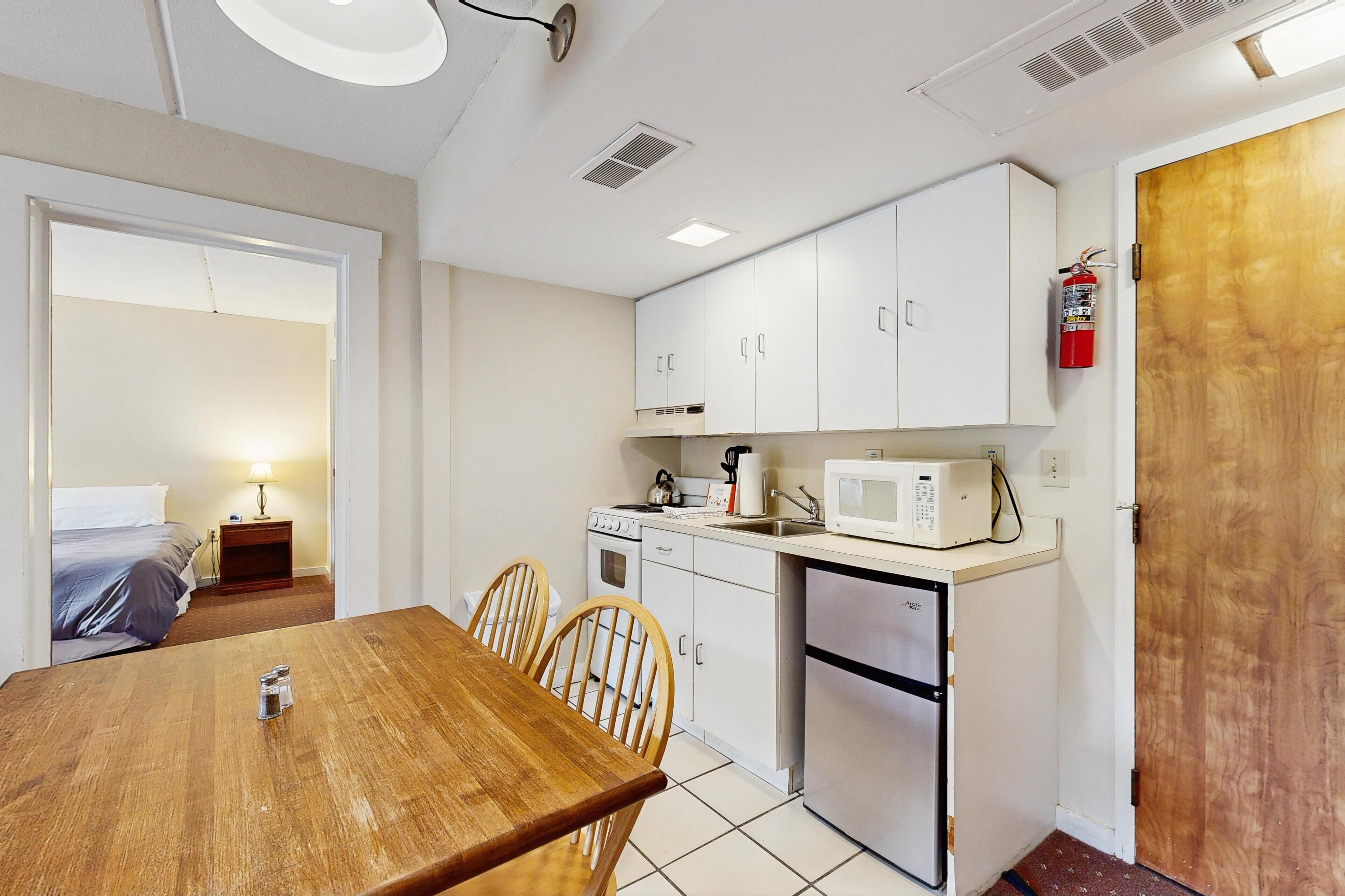 Cocina compacta con armarios blancos, microondas, estufa y refrigerador. Hay una mesa de comedor de madera con sillas.
