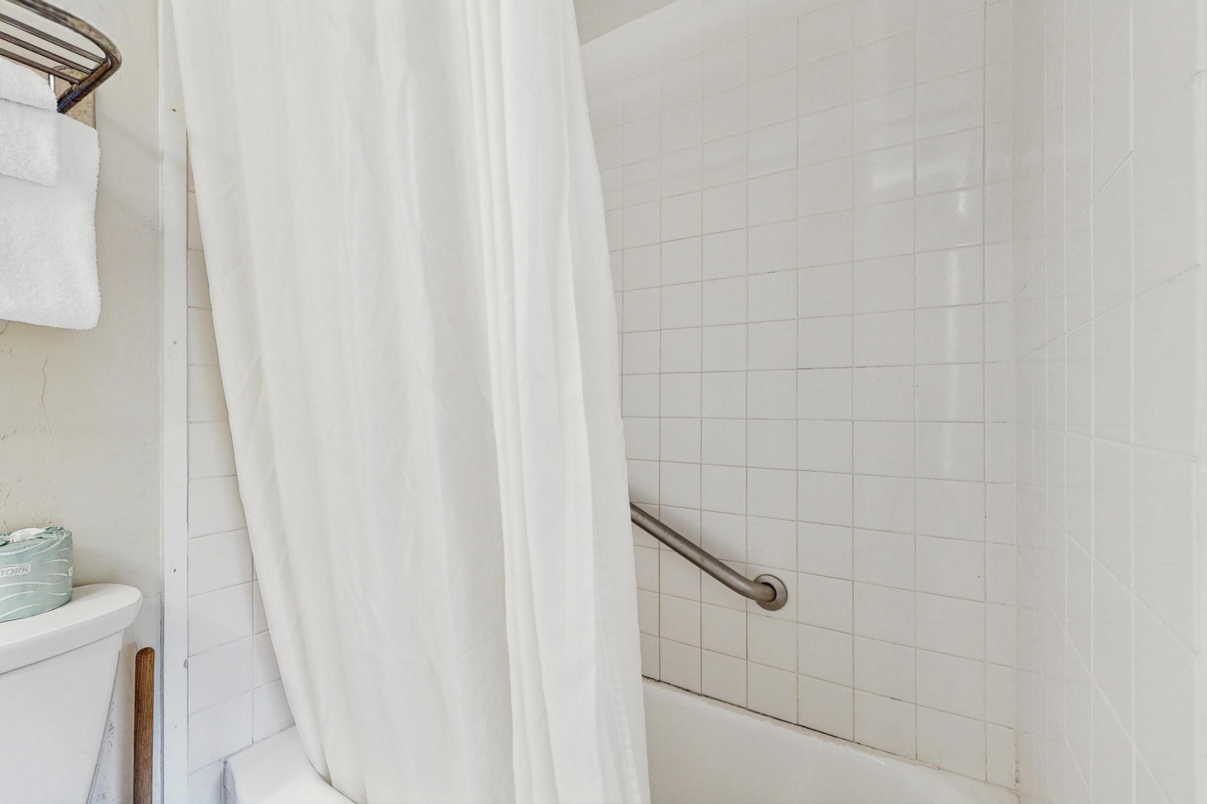 Un baño con ducha, azulejos blancos, una barra de apoyo y un perchero.