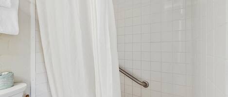 Un baño con ducha, azulejos blancos, una barra de apoyo y un perchero.