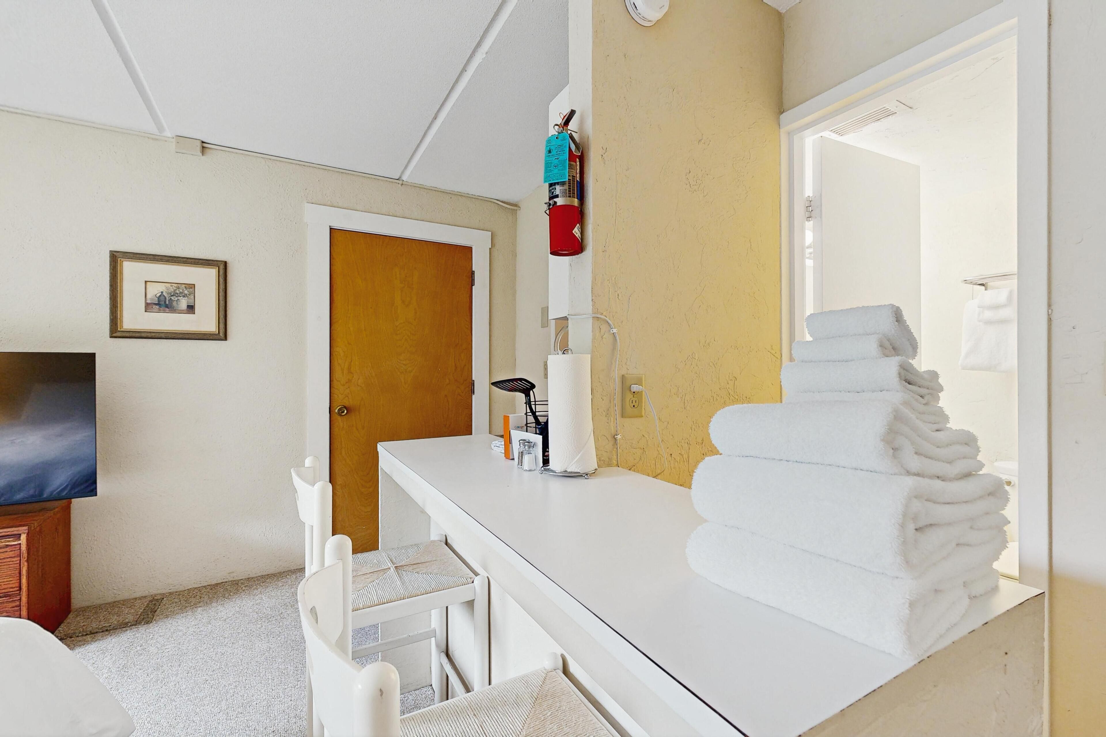 A hotel room with a white vanity, a mirror, a towel stack, a television, and a door.