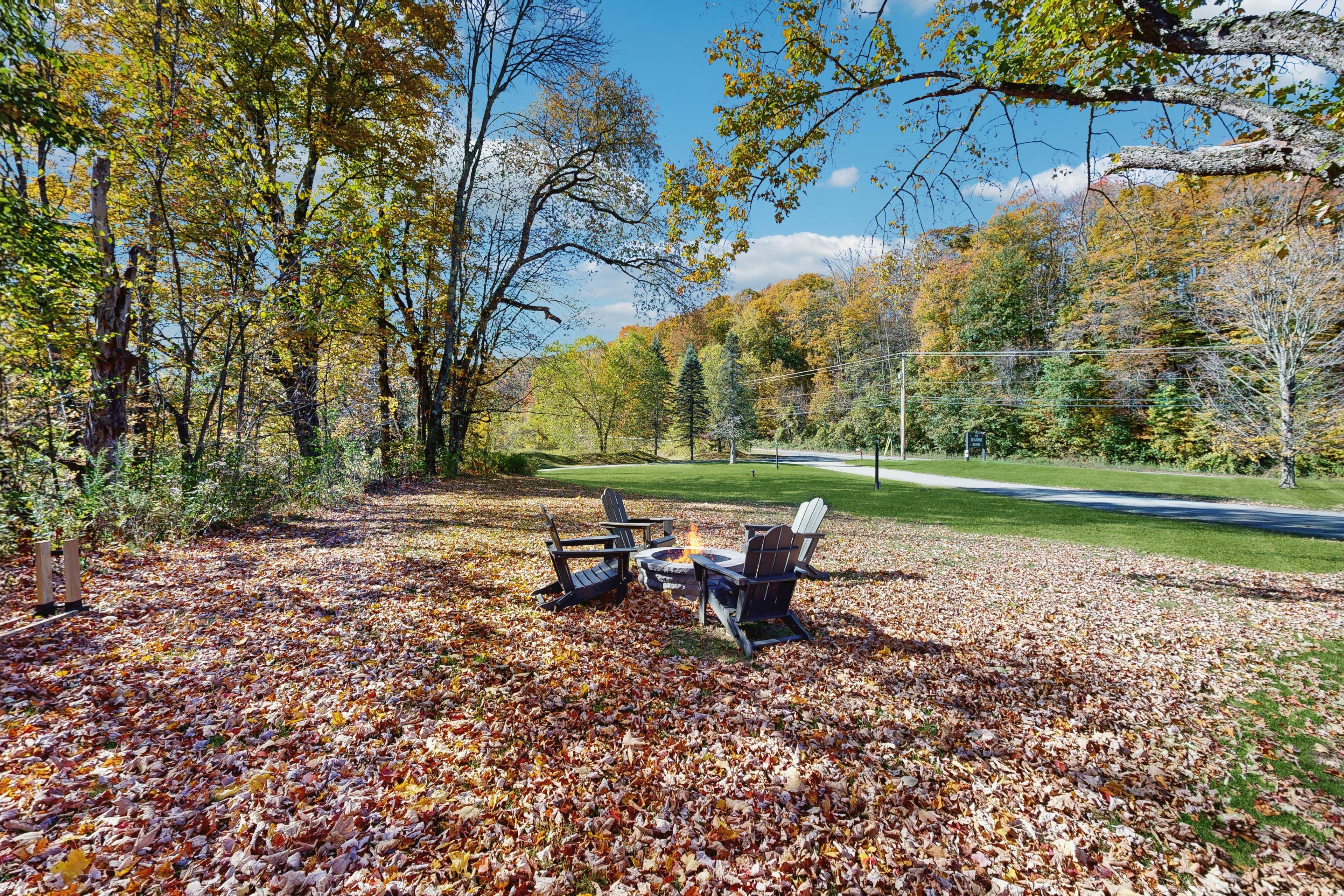 A park with a fire pit and Adirondack chairs on a leaf-covered ground.