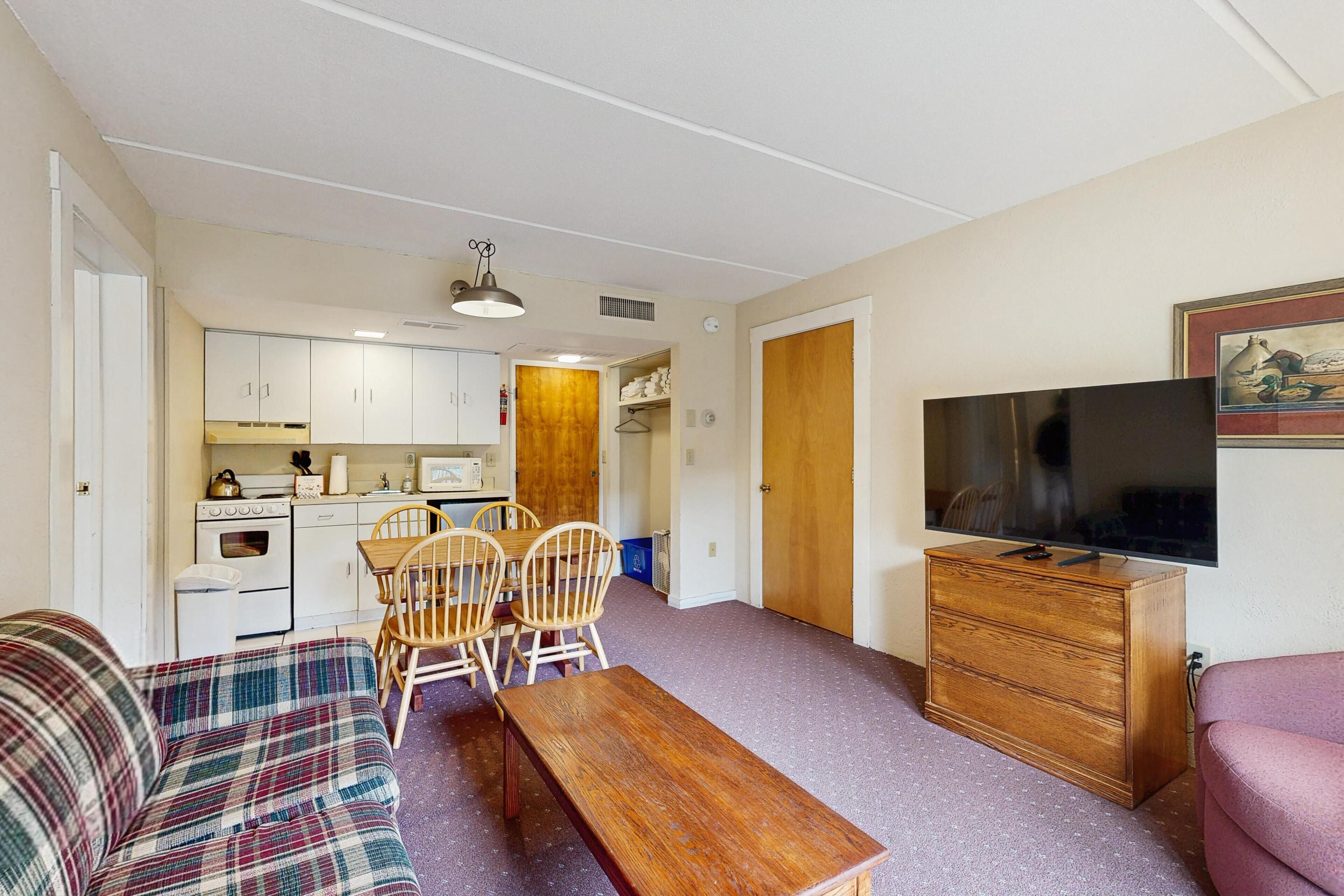 Un espacio habitable compacto con kitchenette, comedor con sillas de madera, sofá, televisor y mueble.