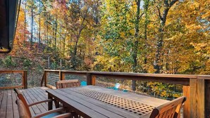 An outdoor wooden deck with a table and chairs, surrounded by trees with autumn foliage. - Endless MTN Escape: Spacious, Hot Tub, MORE GAMES! (Sevierville)
