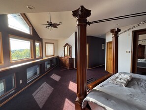 Un dormitorio con una cama grande, un aparador de madera, un espejo, un ventilador de techo y vistas al exterior a través de las ventanas.