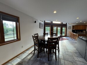 A spacious dining area with a large table and chairs, a kitchen with wooden cabinets, and large windows offering a view of the outdoors.