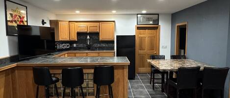 Una cocina moderna con una isla central, taburetes de bar, una mesa de comedor con sillas, armarios de madera y un refrigerador.