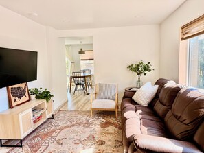 Ein Wohnzimmer mit braunem Ledersofa, einem Fernseher, einem Esstisch mit Stühlen und einem gemusterten Teppich.