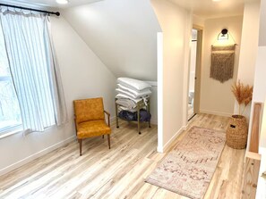 Une pièce avec un plancher en bois, un banc, un lit avec des oreillers, une lampe fixée au mur et un panier en osier.