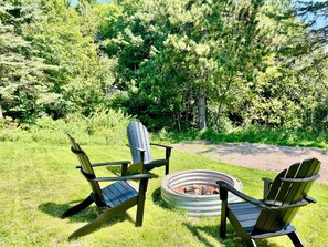 Two Adirondack chairs facing a fire pit in a grassy area with trees in the background.